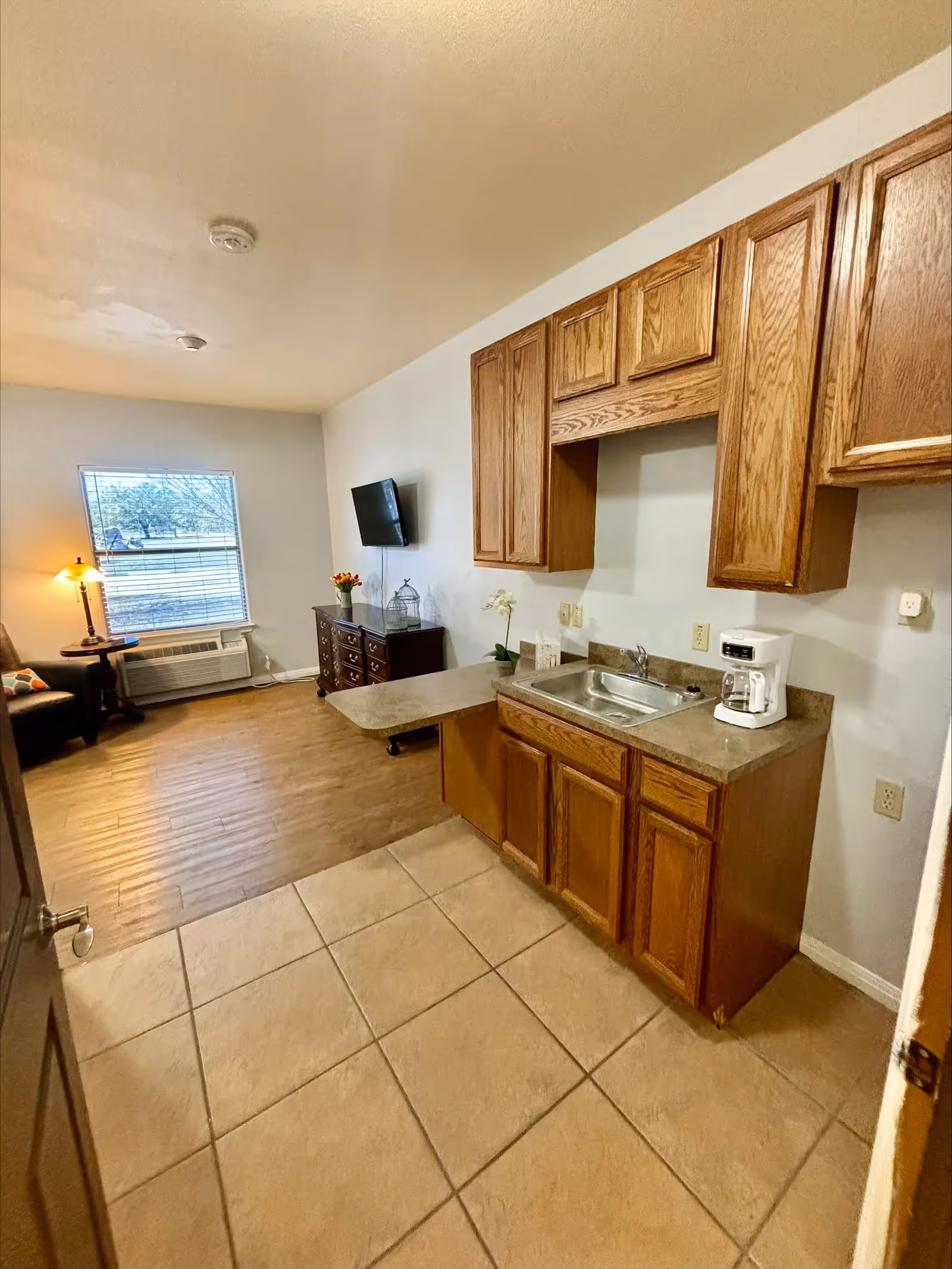 Interior view of a small kitchen area with wooden cabinets, a countertop with a sink and a coffee maker. The kitchen opens into a living area with a window, a wall-mounted TV, a wooden dresser, a side table with a lamp, and a chair. The floor transitions from tiled in the kitchen to wood in the living area.