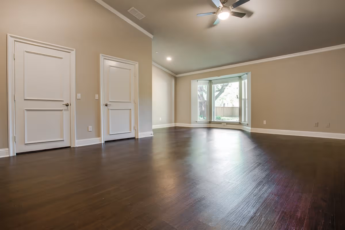 Empty room with dark wooden flooring, beige walls, two white doors on the left wall, a ceiling fan with light, and a large bay window showing a view of trees outside.