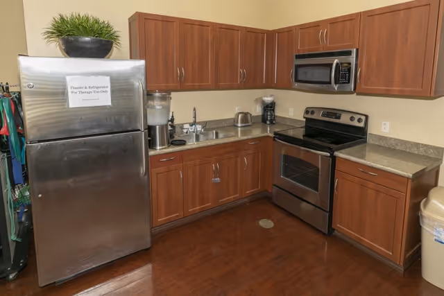 A kitchen area with wooden cabinets, a stainless steel refrigerator with a sign that reads 'Freezer & Refrigerator For Therapy Use Only,' a blender, toaster, coffee maker, stainless steel sink, electric stove with oven, microwave, and a trash can. The floor is wooden and there is a small plant on top of the refrigerator.