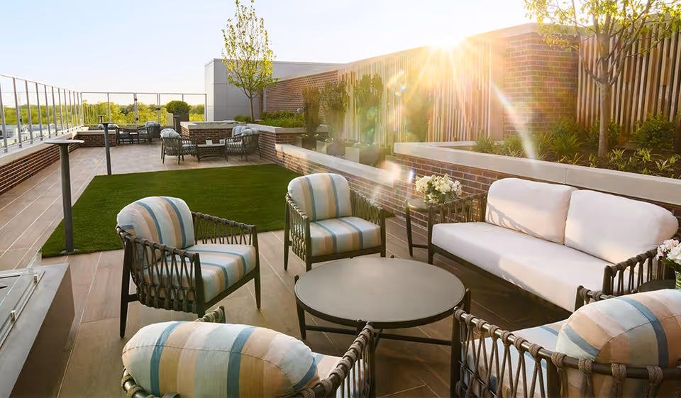 Rooftop outdoor lounge with cushioned chairs, a sofa and a round table beside a patch of turf and planters in bright sunlight.