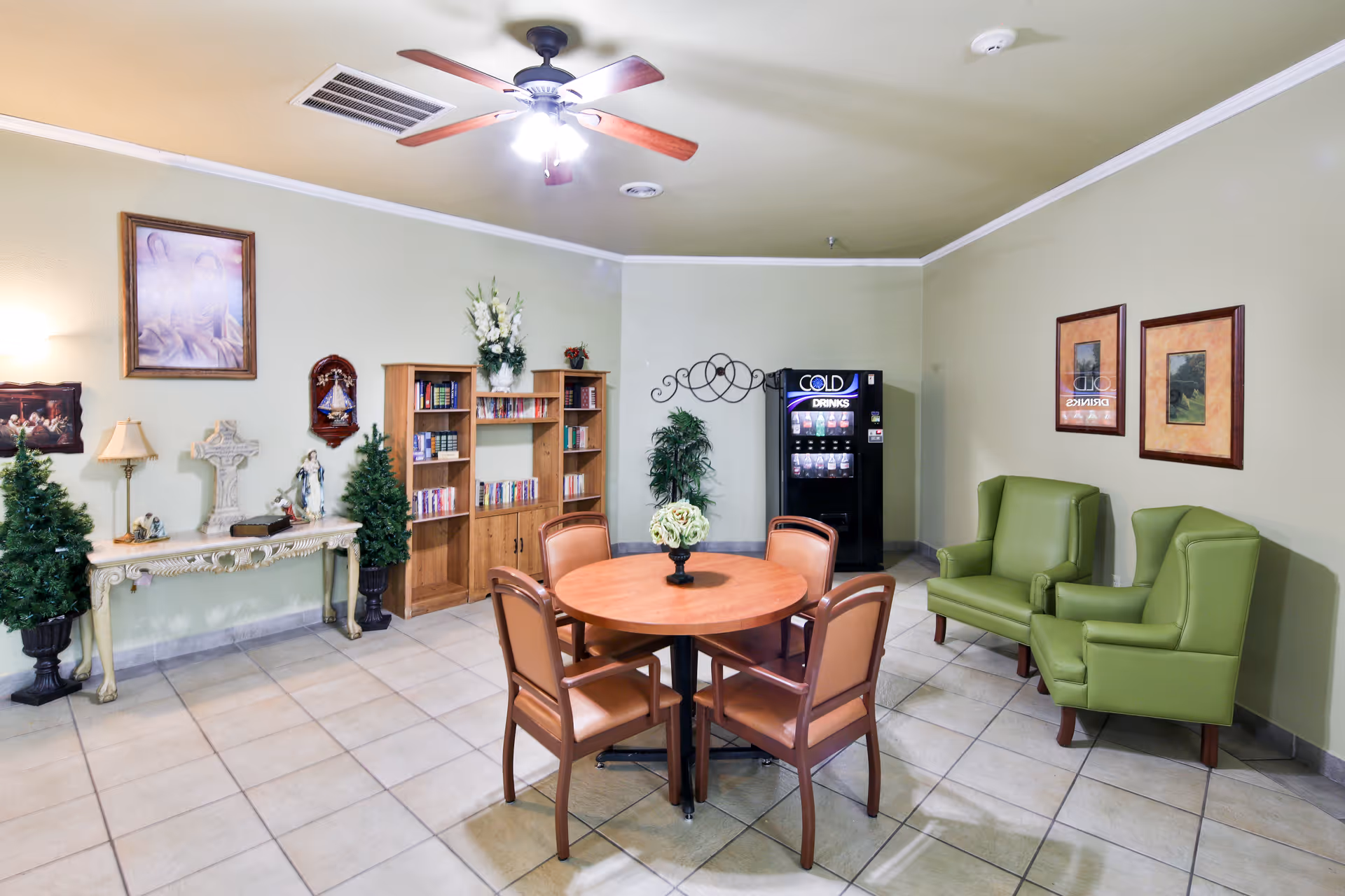 A cozy common area with a round wooden table surrounded by four chairs in the center. Two green armchairs are positioned against the right wall. A black vending machine labeled 'Cold Drinks' stands in the corner. On the left side, there is a decorative console table with a lamp, statues, and two small artificial pine trees. A wooden bookshelf filled with books and decorative items is also visible. The walls are painted light green, adorned with framed pictures and a ceiling fan with wooden blades is mounted above.