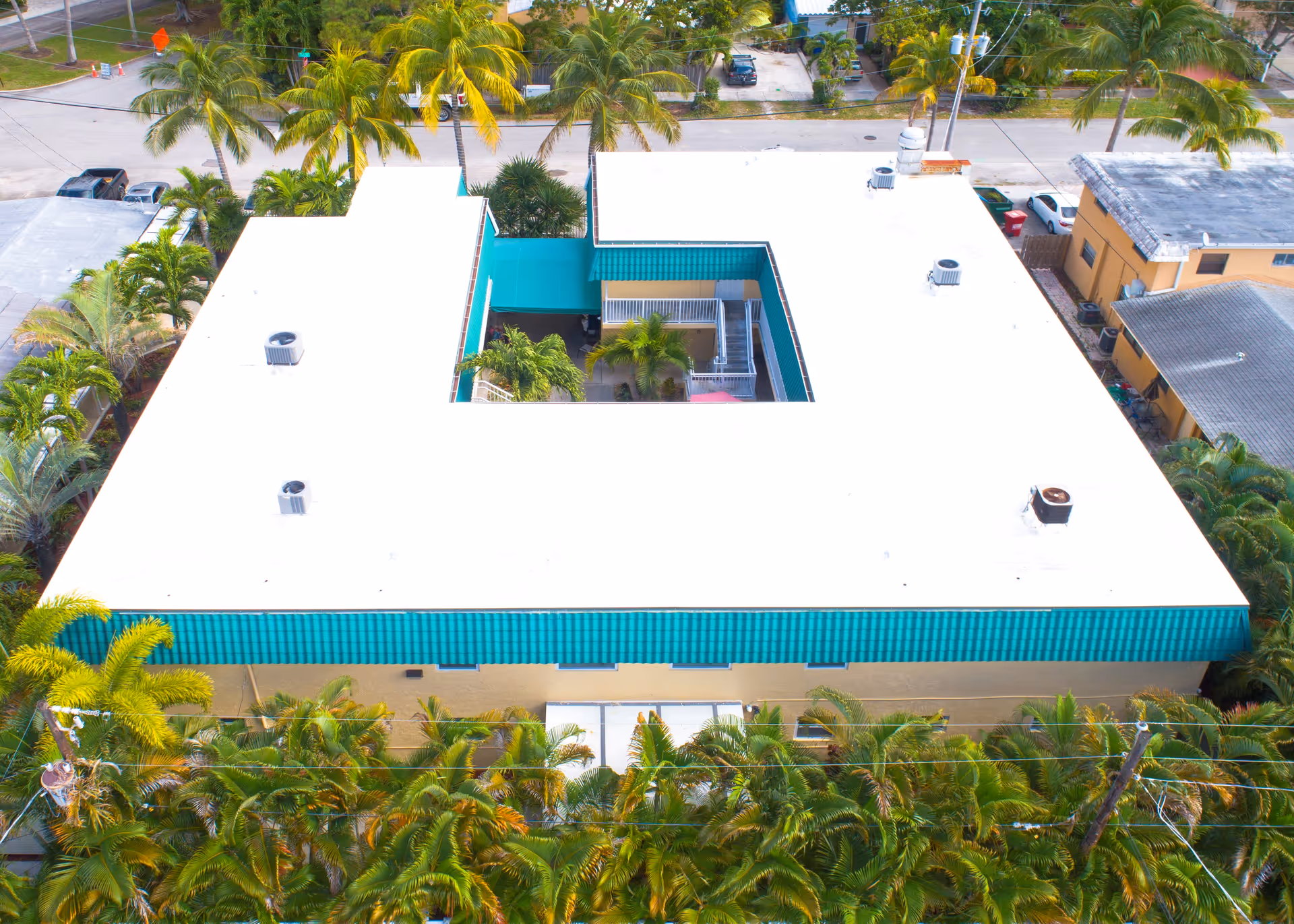Aerial view of a U-shaped building with a white roof and teal trim, surrounded by palm trees and greenery. The building has an open courtyard in the center with visible stairs and plants. Several cars are parked on the street nearby.