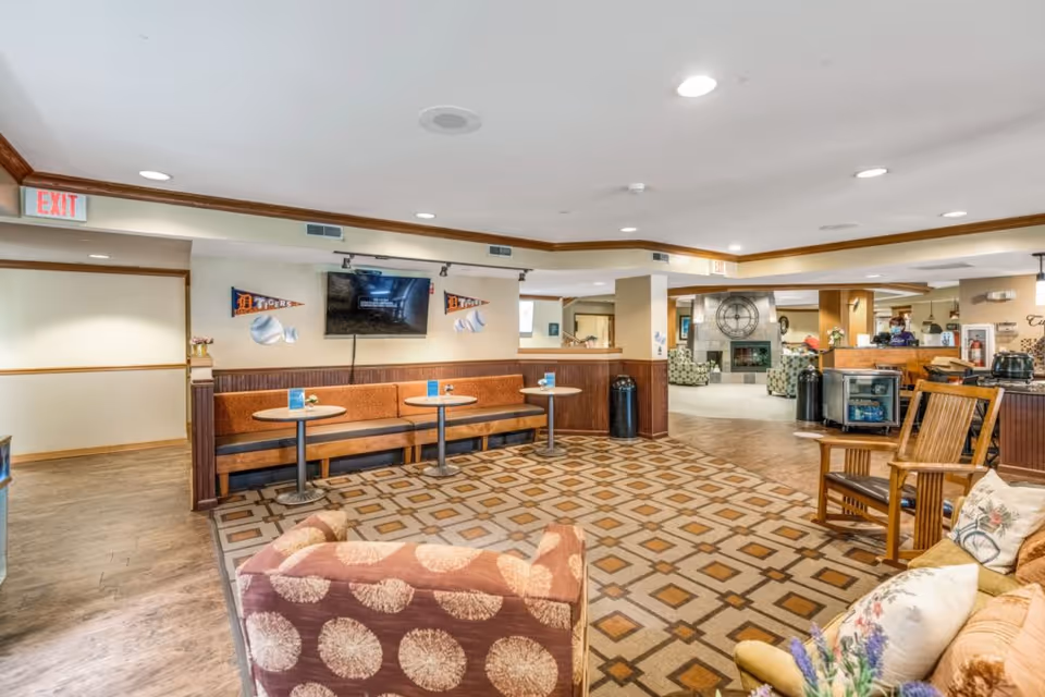 Open communal lounge with benches, small tables, upholstered chairs, a wall-mounted TV and a fireplace in the background.