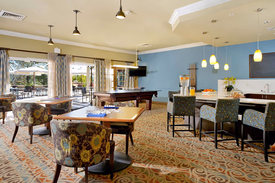 A bright and spacious common area with patterned carpet, several tables with floral upholstered chairs, a pool table, and a kitchen counter with bar stools. Large windows and glass doors open to an outdoor patio with tables and umbrellas. The walls are painted in light yellow and blue, and pendant lights hang from the ceiling.