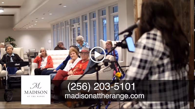 A group of elderly people seated in a well-lit common area with large windows, listening to a woman speaking or singing into a microphone. The setting appears to be a senior living facility lounge or common room.