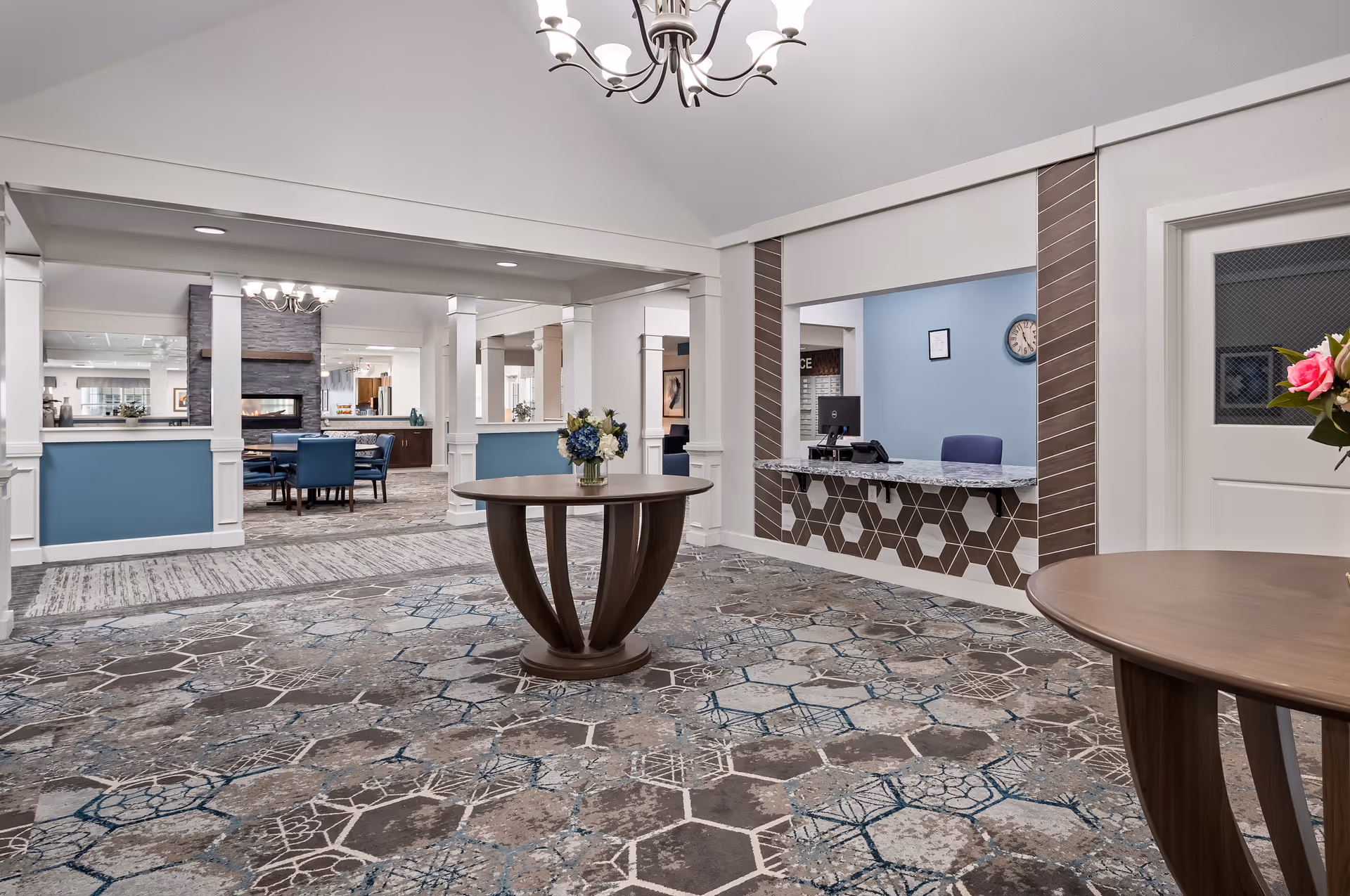 Interior view of a senior living facility lobby area with patterned carpet, two round wooden tables with flower arrangements, a reception desk with a computer, and a seating area with blue chairs and a fireplace in the background.
