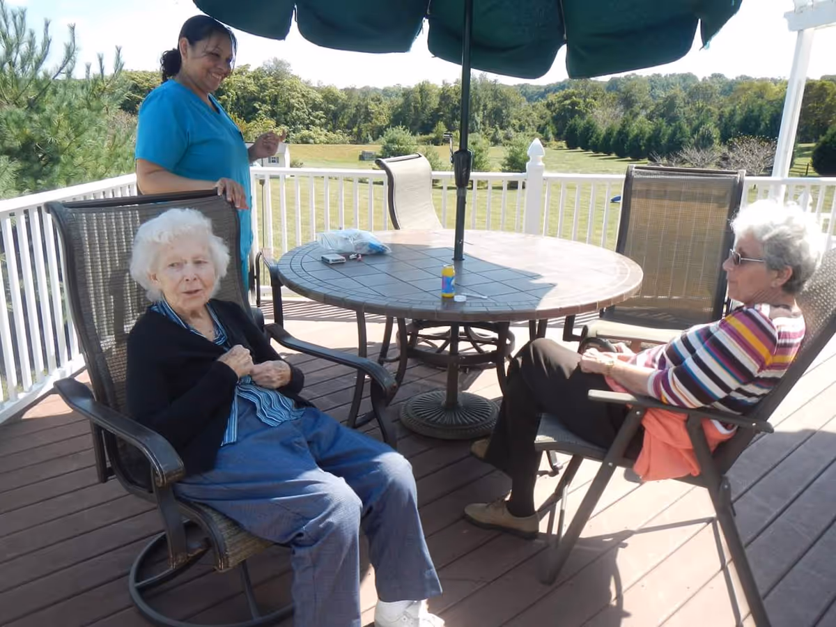 Two elderly women sit at a round patio table under an umbrella on a deck while a caregiver stands nearby and trees and lawn are visible beyond the railing.