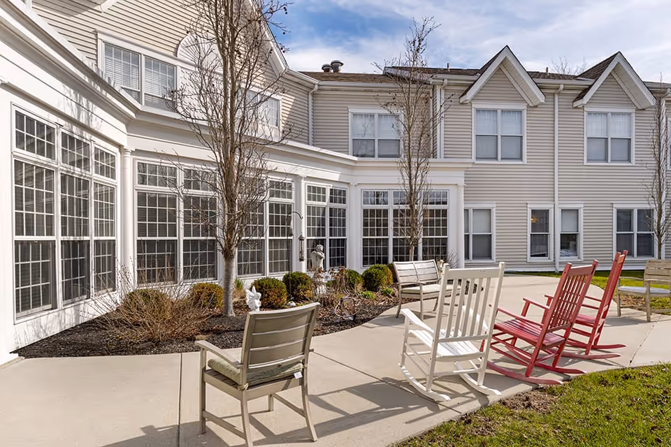 Outdoor patio area at Brookdale Kenwood featuring several rocking chairs and benches arranged on a concrete walkway. The patio is adjacent to a beige building with large windows and some small trees and shrubs in landscaped beds.