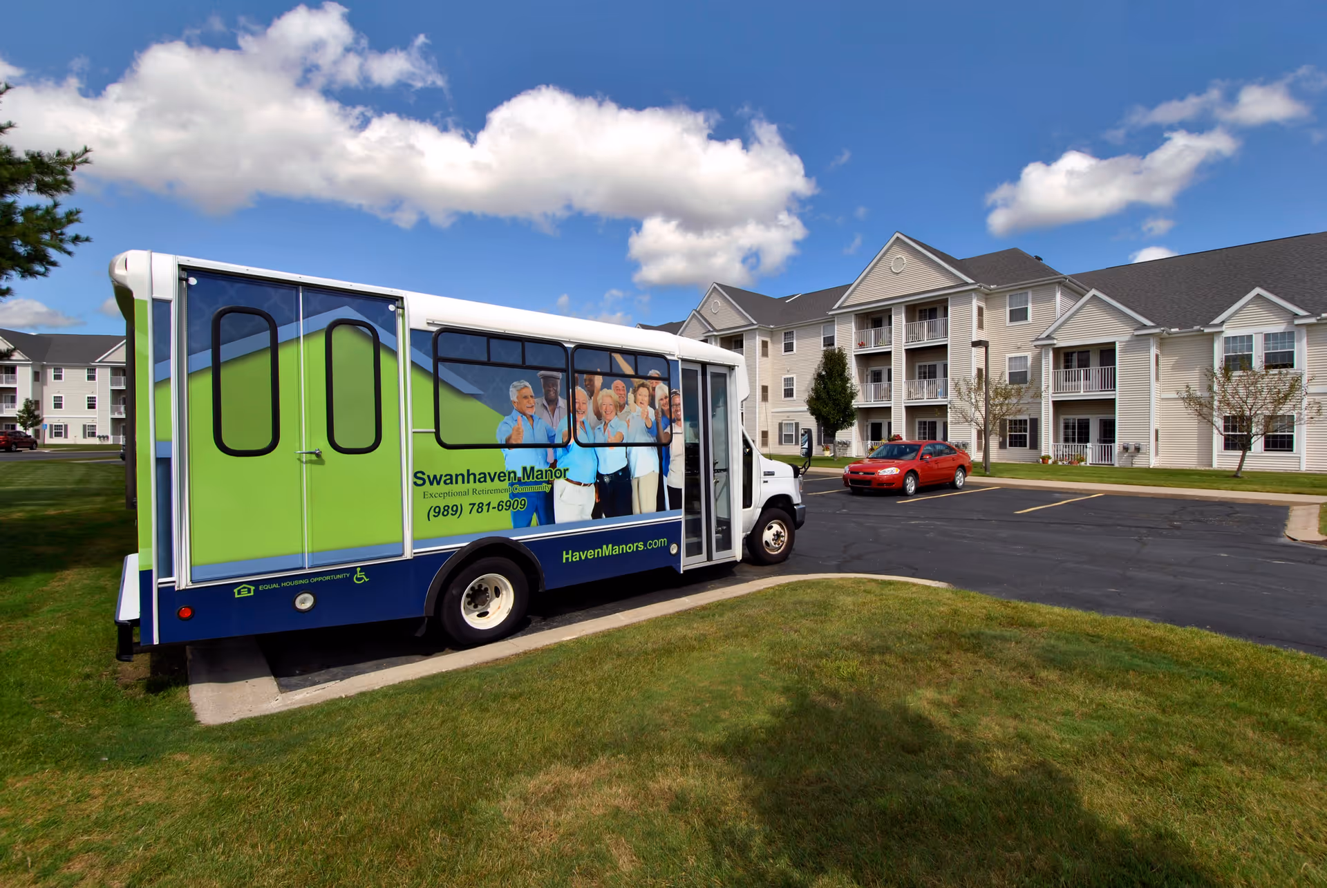 A shuttle bus parked on a driveway in front of a multi-story retirement community building under a blue sky with scattered clouds. The bus has a green and blue design with images of smiling elderly people and text advertising Swanhaven Manor Retirement Community with a phone number and website. A red car is parked near the building.