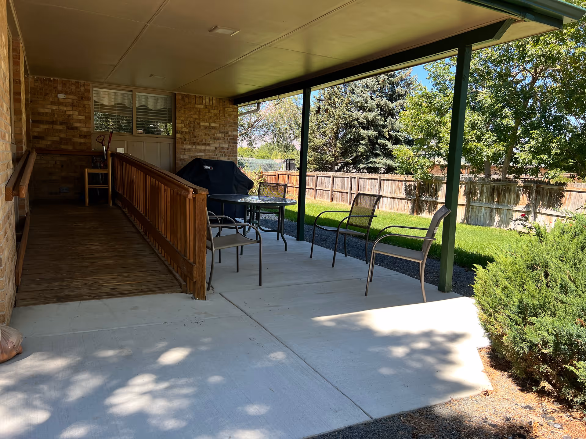 Covered outdoor patio area with a wooden ramp leading to a door, a round glass table with four metal chairs, a barbecue grill, and a fenced backyard with green grass and trees.