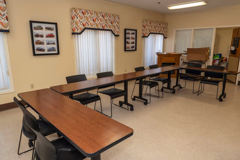 A meeting or activity room with tables arranged in a U-shape and black chairs around them. The room has beige walls with two windows covered by patterned valances and sheer curtains. There are framed pictures of vintage cars on the walls, and a wooden cabinet with a box and other items on top is visible in the background.