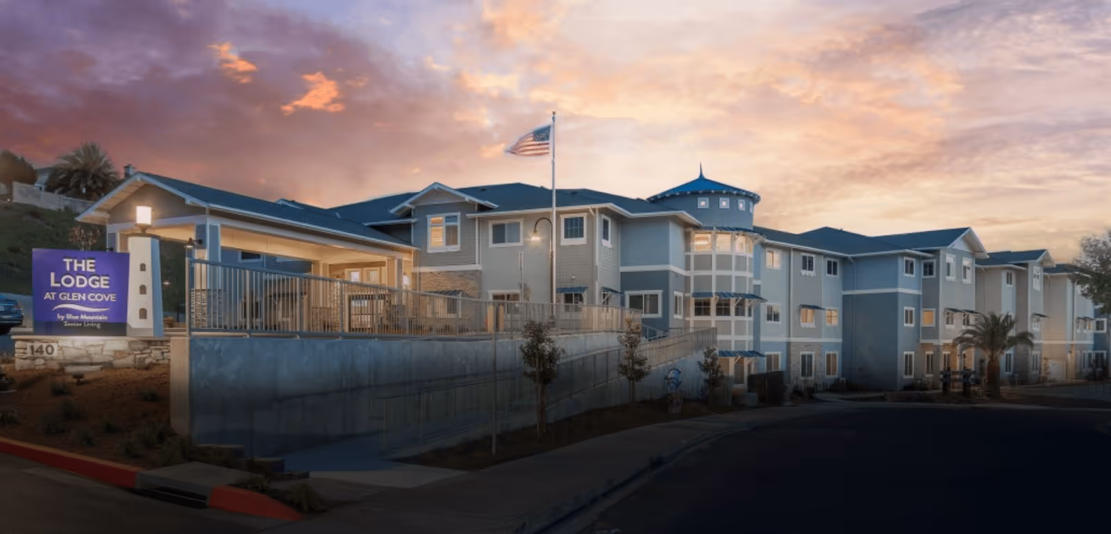 Exterior view of The Lodge at Glen Cove senior living facility during sunset, showing a large multi-story building with a flagpole flying the American flag and a sign at the entrance.