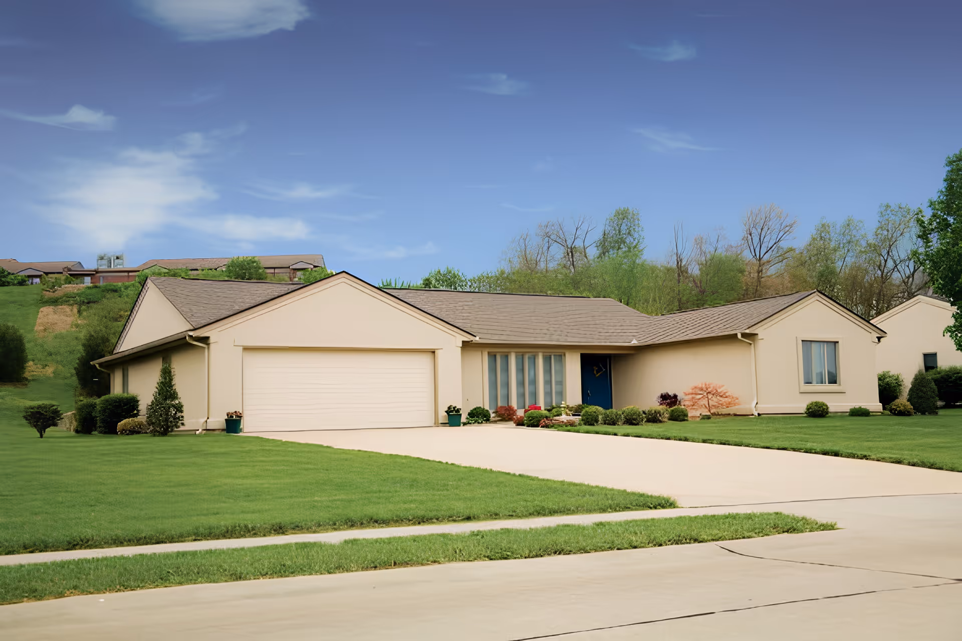 Single-story beige house with a two-car garage, a blue front door, and a well-maintained lawn with shrubs and small trees under a clear blue sky.