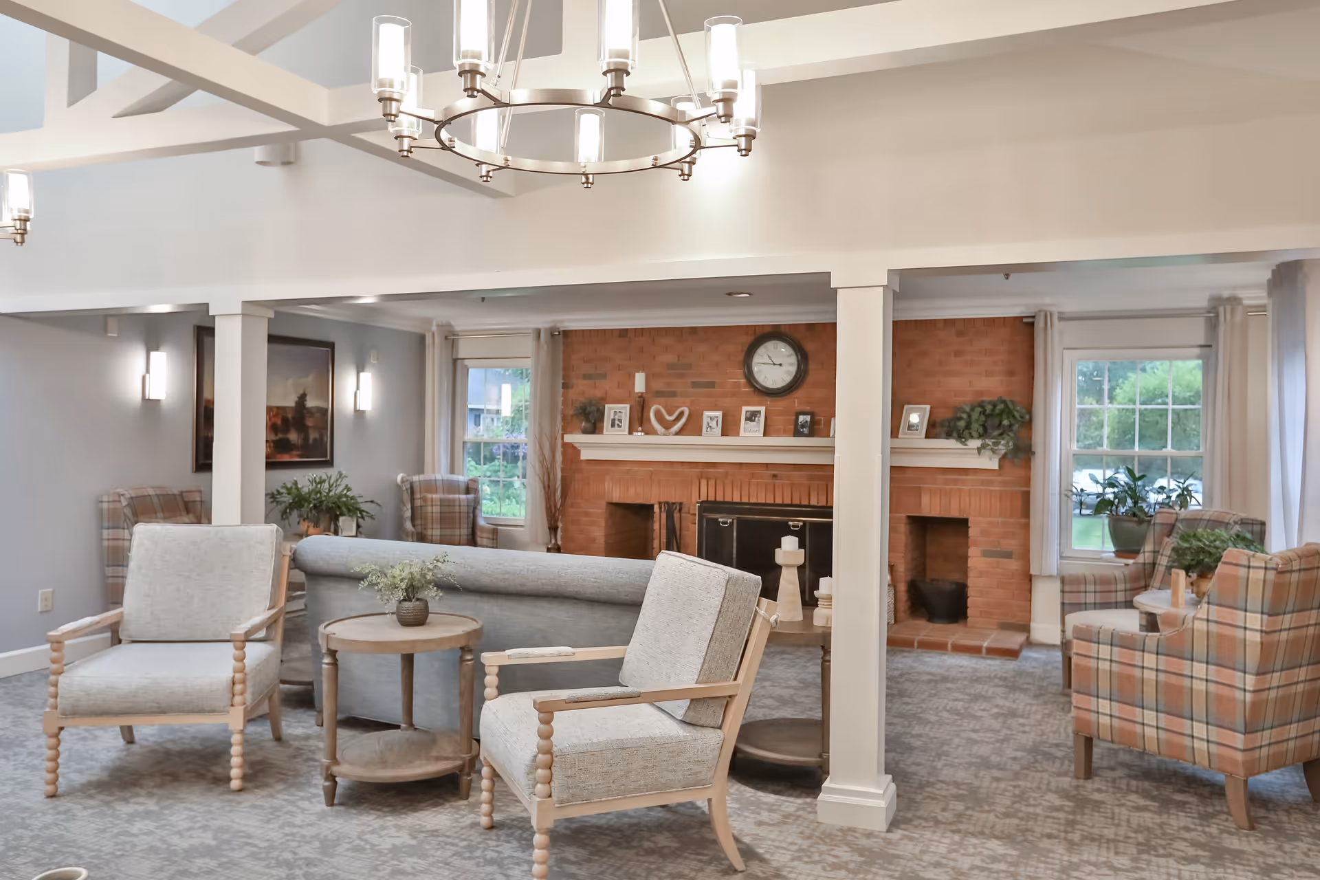 A cozy senior living facility common area with a brick fireplace, a clock above the mantel, framed photos, and plants. The room features comfortable seating including a gray sofa, two light gray armchairs with wooden frames, and two plaid armchairs. There are windows with light curtains letting in natural light, and a modern chandelier hanging from the ceiling.