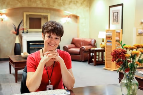 A smiling woman with short brown hair wearing a red shirt and a name badge sits at a desk in a warmly decorated room. Behind her, there is a fireplace, a brown leather armchair, a wooden side table, and a rack with magazines or brochures. A vase with colorful flowers is on the desk in the foreground.