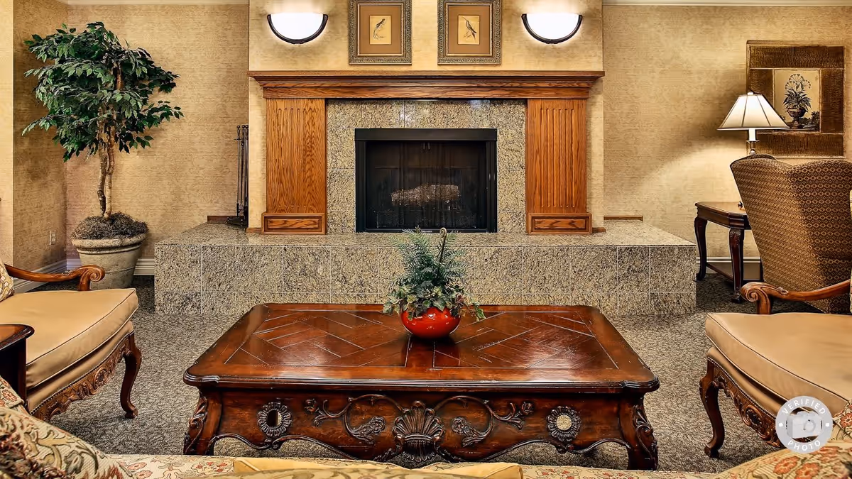 A cozy living room area featuring a stone fireplace with a wooden mantle, two wall-mounted lights above it, and framed bird artwork. In front of the fireplace is a polished wooden coffee table with a red vase containing green plants. The room has upholstered armchairs and a potted plant on the left side, with a table and lamp on the right side.