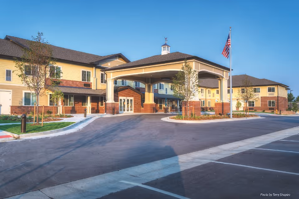 Exterior view of a large assisted living and memory care facility building with a covered entrance, multiple windows, small trees, and an American flag on a flagpole in front. The sky is clear and blue.