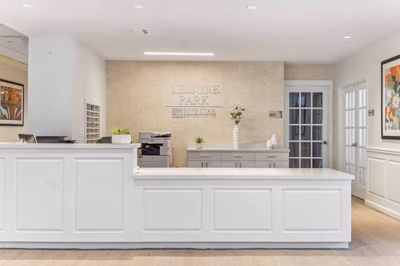 Reception area of Leisure Park Five Star Senior Living facility with a white front desk, a printer, decorative plants, and framed artwork on the walls.