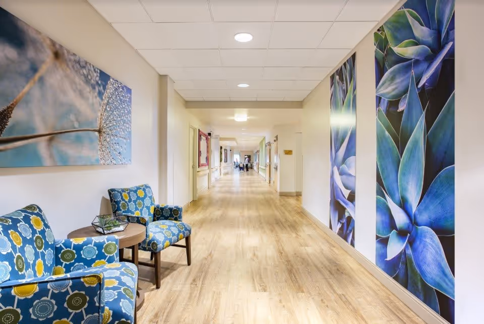 A long, well-lit hallway with light wood flooring and white walls. On the left side, there are two blue patterned armchairs with a small round table between them, decorated with a small plant. The walls feature large, colorful botanical artwork, including close-up images of flowers and plants. At the far end of the hallway, a few people are visible in the distance.