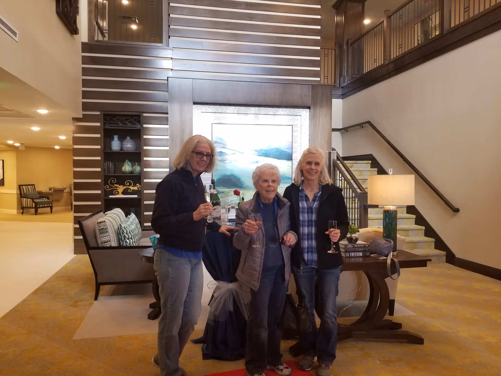 Three women holding wine glasses stand in a lobby area with seating, decorative shelving, a table, and a staircase behind them.