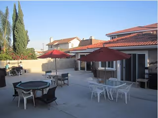 Outdoor patio area with several tables and chairs, red umbrellas, and a single-story building with a red tile roof.