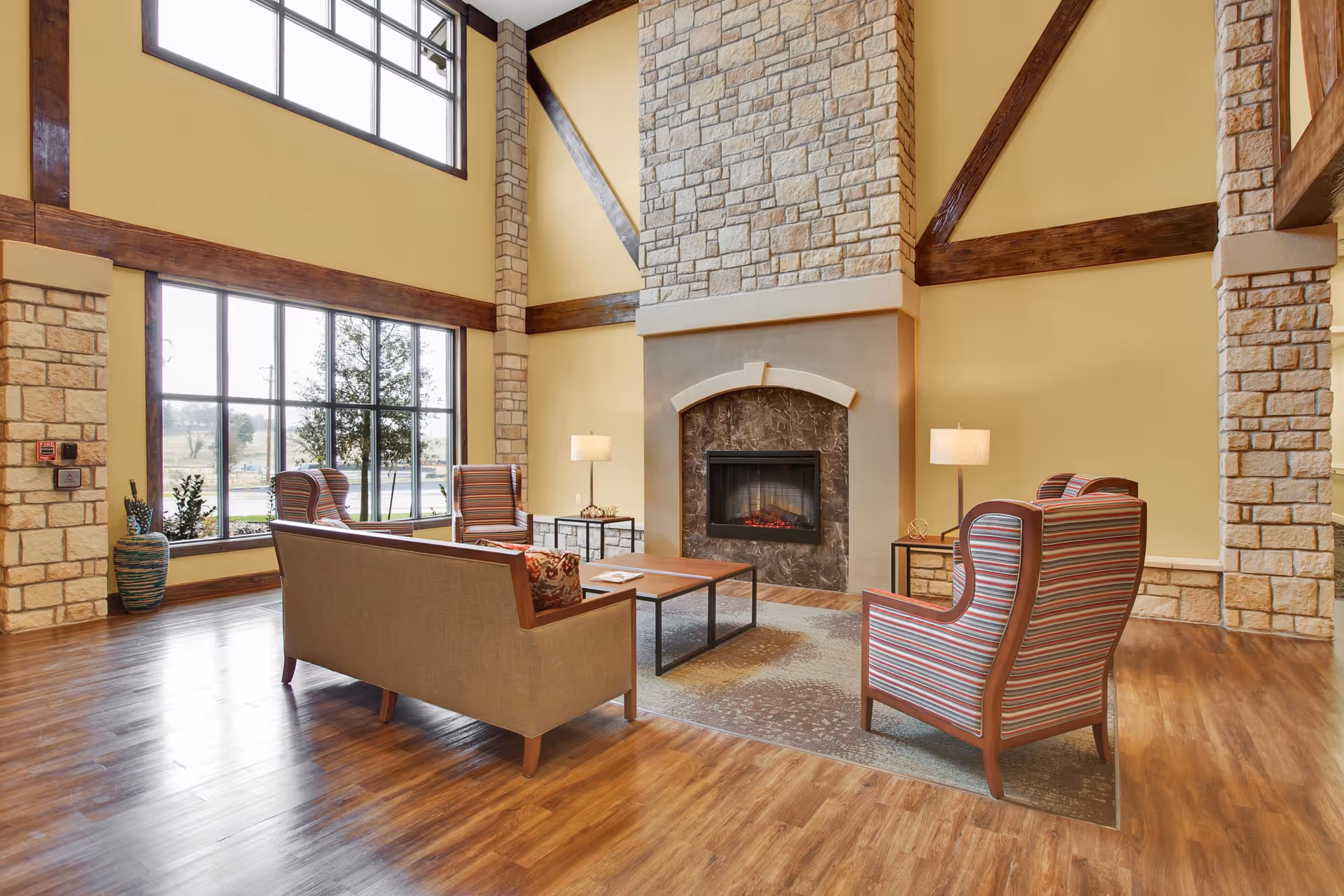 A spacious living room with high ceilings featuring large windows, a stone fireplace, wooden beams, and wood flooring. The room is furnished with a beige sofa, three striped armchairs, two side tables with lamps, and a coffee table on a patterned rug.