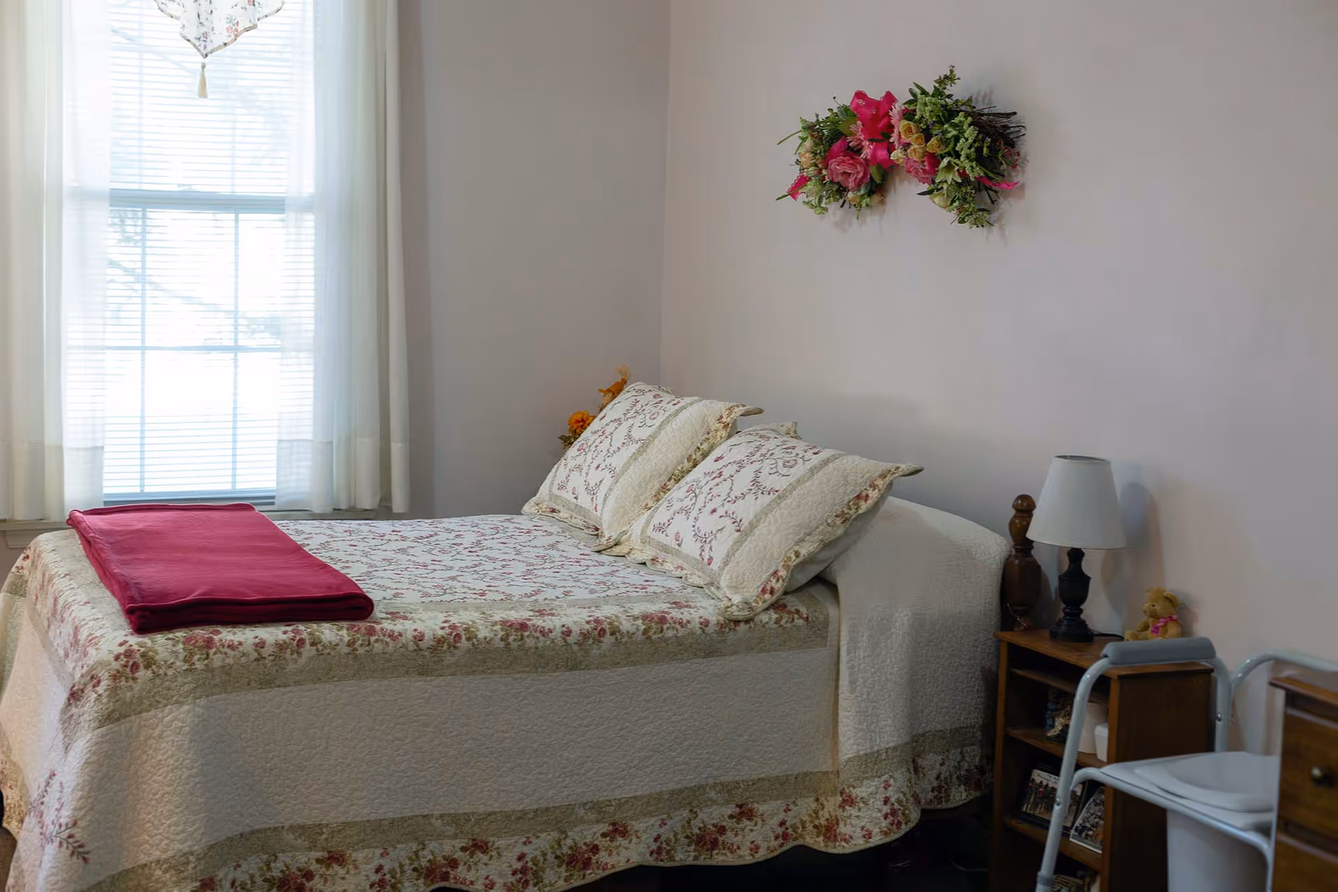 Bright bedroom with a floral quilted bed by a curtained window, a nightstand with a lamp, and a walker.