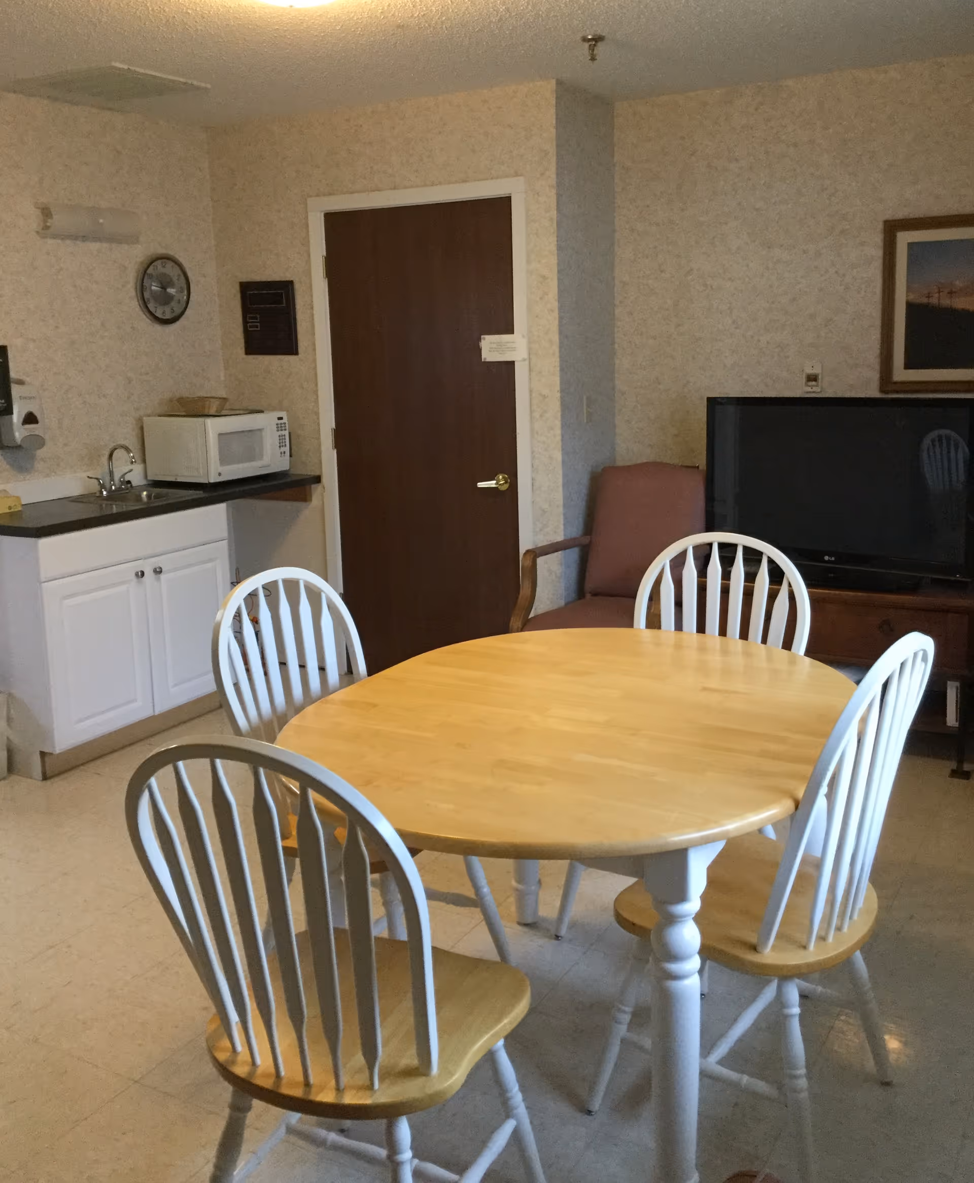 Small dining area with a round wooden table and four chairs in a room with a kitchenette, TV, and armchair.