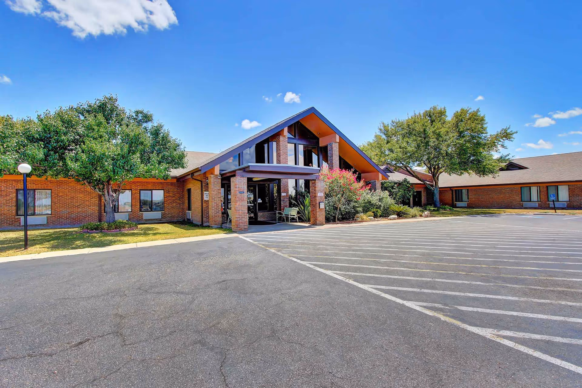 Exterior view of Caraday of Corpus Christi Assisted Living facility showing a brick building with a peaked roof entrance, surrounded by trees and landscaping under a clear blue sky.