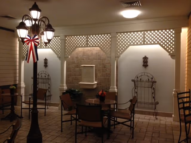 Indoor seating area with a round table and four chairs, decorative wrought iron wall hangings, a tiled wall with a small fountain, and a lamp post adorned with a red, white, and blue ribbon. The space has a tiled floor and white lattice trim along the upper walls.