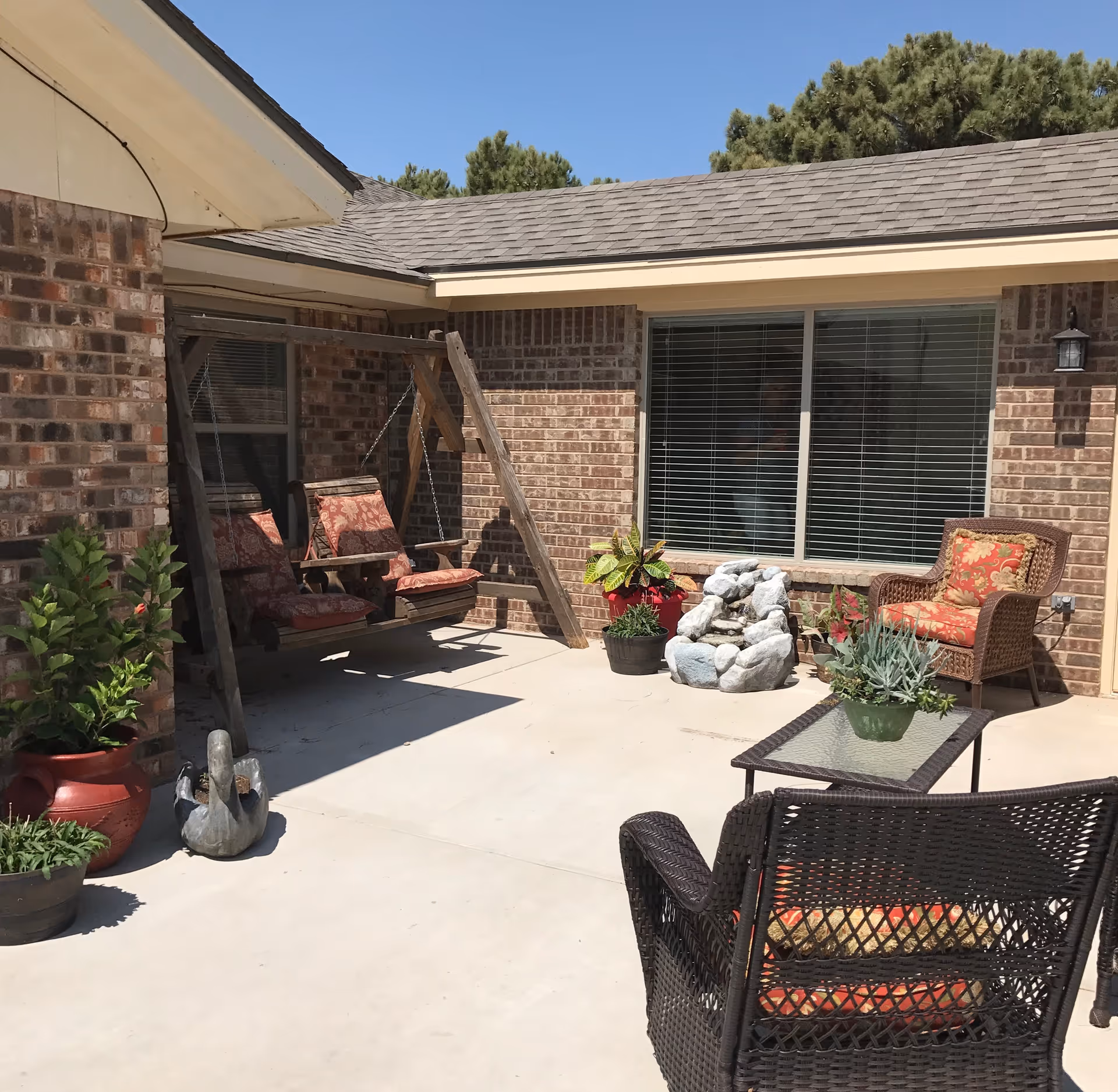 Sunny brick patio with wicker chairs, a cushioned wooden swing, potted plants and a small rock fountain by a window.