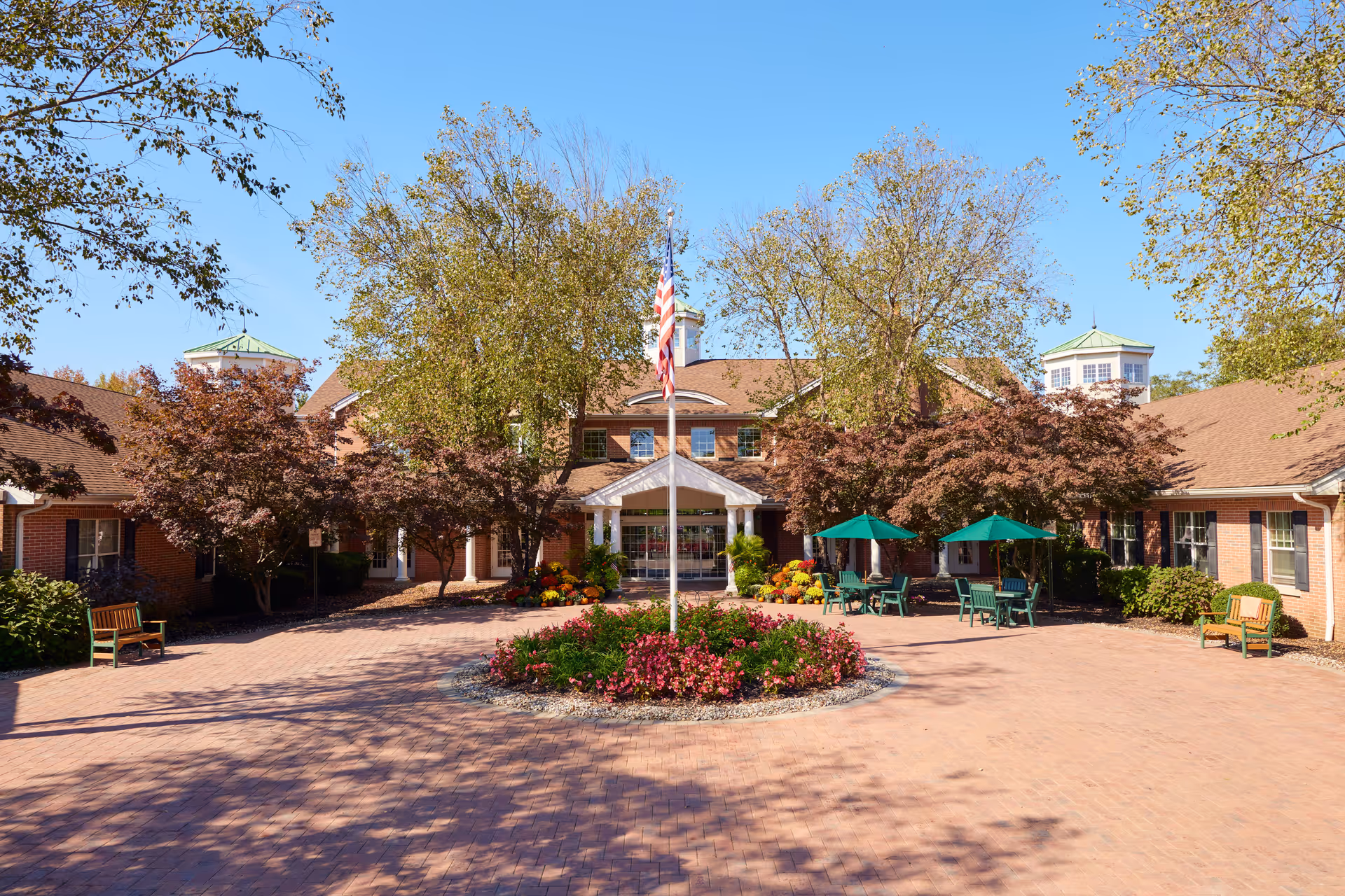 Front exterior view of Brandywine Colts Neck by Monarch facility with a brick paved circular driveway, a flagpole with an American flag in the center surrounded by flowers, trees with autumn foliage, green patio umbrellas with tables and chairs, and benches on either side.