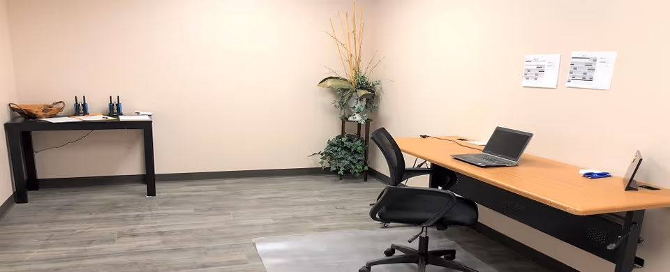 A small office room with a wooden desk holding a laptop, papers, and pens. There is a black office chair on wheels in front of the desk. Against the left wall is a smaller black table with two walkie-talkies and some papers. A plant with green leaves and tall decorative grass is placed in the corner. The floor has a gray wood-like finish and the walls are beige with two sheets of paper pinned on the right wall.