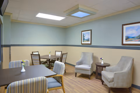 Small communal dining and seating area with tables, chairs, armchairs, framed artwork, and overhead lighting.