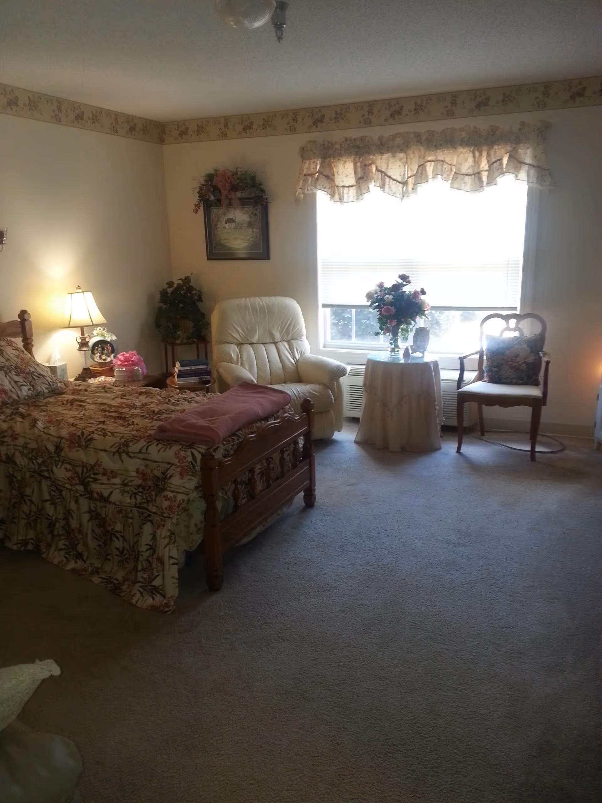 A cozy bedroom with a wooden bed covered in floral bedding, a white recliner chair, a small round table with a floral arrangement, and a wooden chair with a cushion. The room has a large window with a floral valance, beige walls with floral border wallpaper near the ceiling, and carpeted floor.