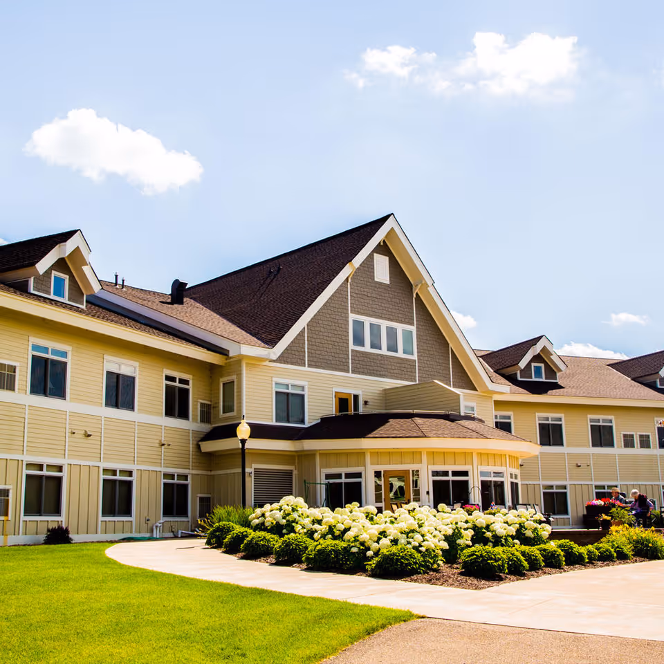 Front exterior of a large two-story assisted living building with a landscaped entrance and beds of white flowering shrubs.