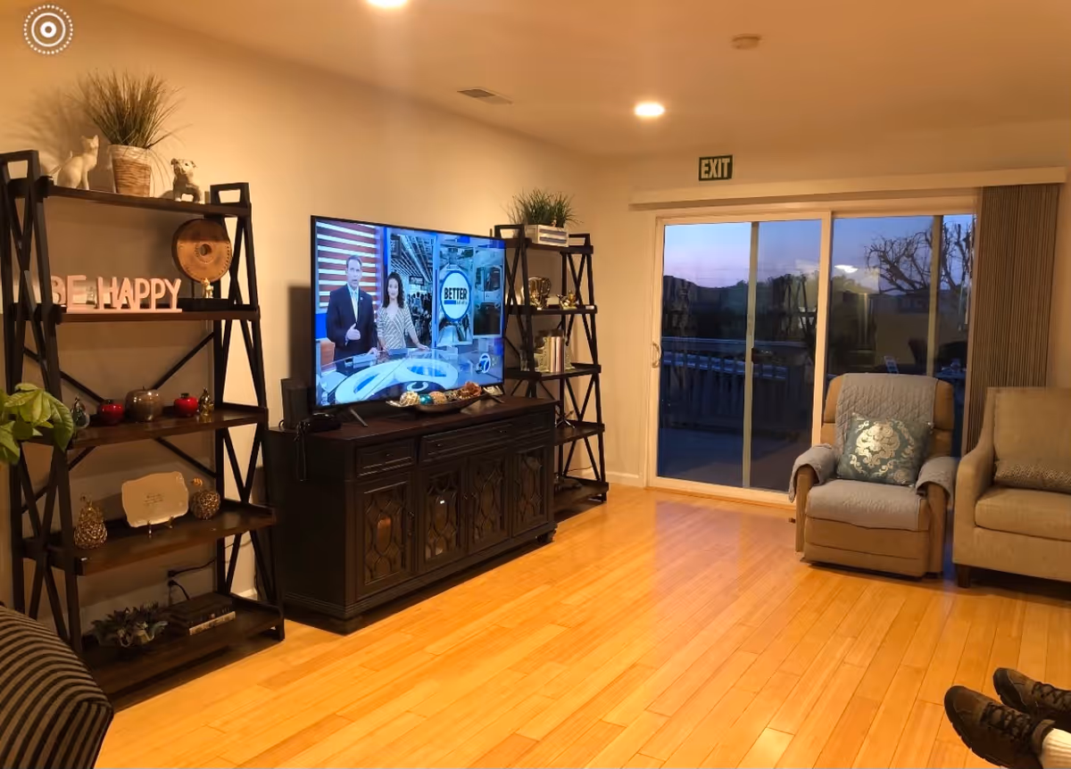 A cozy living room with wooden flooring, a TV on a dark wooden cabinet displaying a news program, two black shelving units with decorative items including plants, figurines, and a 'BE HAPPY' sign. There is a sliding glass door leading to an outdoor area with a view of trees and hills at dusk. Two armchairs are positioned near the door, one with a blue quilted cover and a decorative pillow.