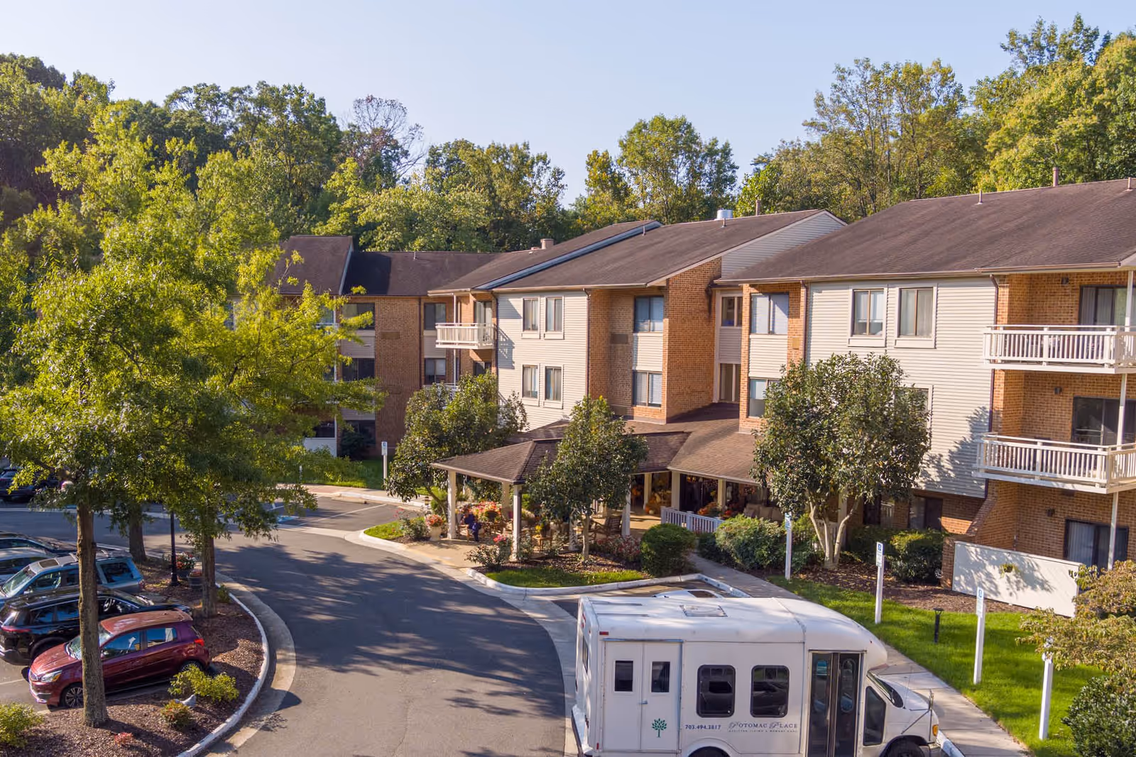 Exterior view of Potomac Place Assisted Living and Memory Care facility showing a three-story building with balconies, surrounded by trees and greenery. A driveway curves in front of the entrance where a white shuttle bus is parked. Several cars are parked along the driveway under the shade of trees.