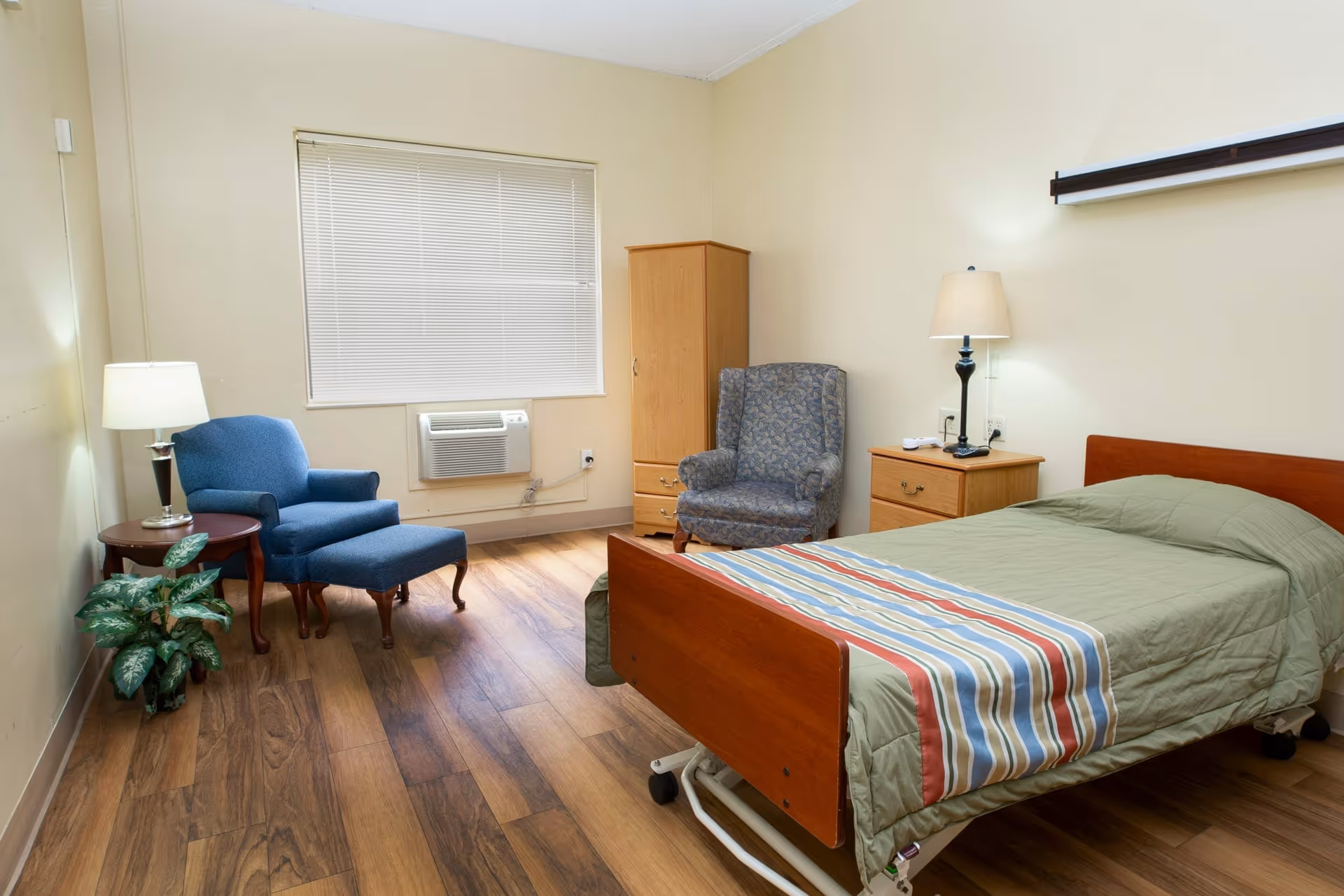A senior living facility bedroom with a single bed covered in a green quilt and a striped blanket. The room has wooden flooring, a wooden nightstand with a lamp, a wooden wardrobe, a blue armchair with an ottoman, a patterned armchair, a small round wooden table with a lamp, and a potted plant near the wall. A window with closed blinds and an air conditioning unit is visible on the wall.