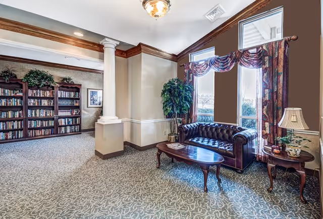 A cozy interior common area with a dark leather tufted sofa, wooden coffee table, and side table with a lamp and plant. Large windows with patterned curtains let in natural light. In the background, there is a bookshelf filled with books and decorative greenery on top. The room features carpeted flooring, crown molding, and a decorative column.