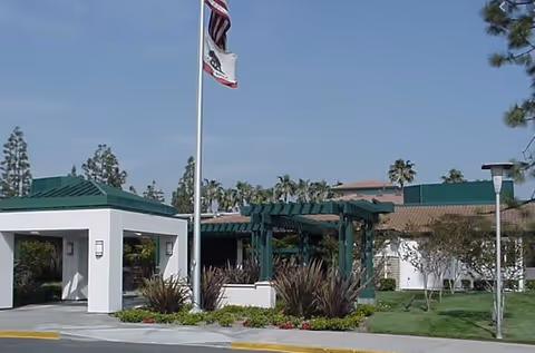 Exterior view of a senior living community building with a covered entrance, flagpoles with American and California state flags, landscaped greenery, and a clear blue sky.