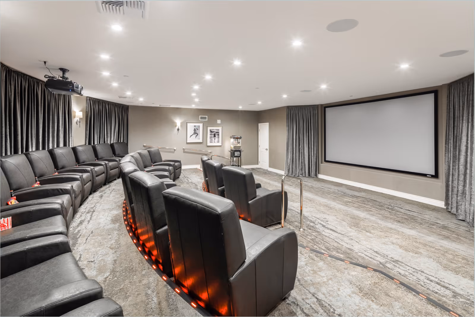 A modern senior living facility theater room with two rows of black leather recliner chairs arranged in a curved layout facing a large blank projection screen. The room has gray walls, carpeted floor, ceiling lights, and dark curtains on the windows. A popcorn machine is visible in the back corner.