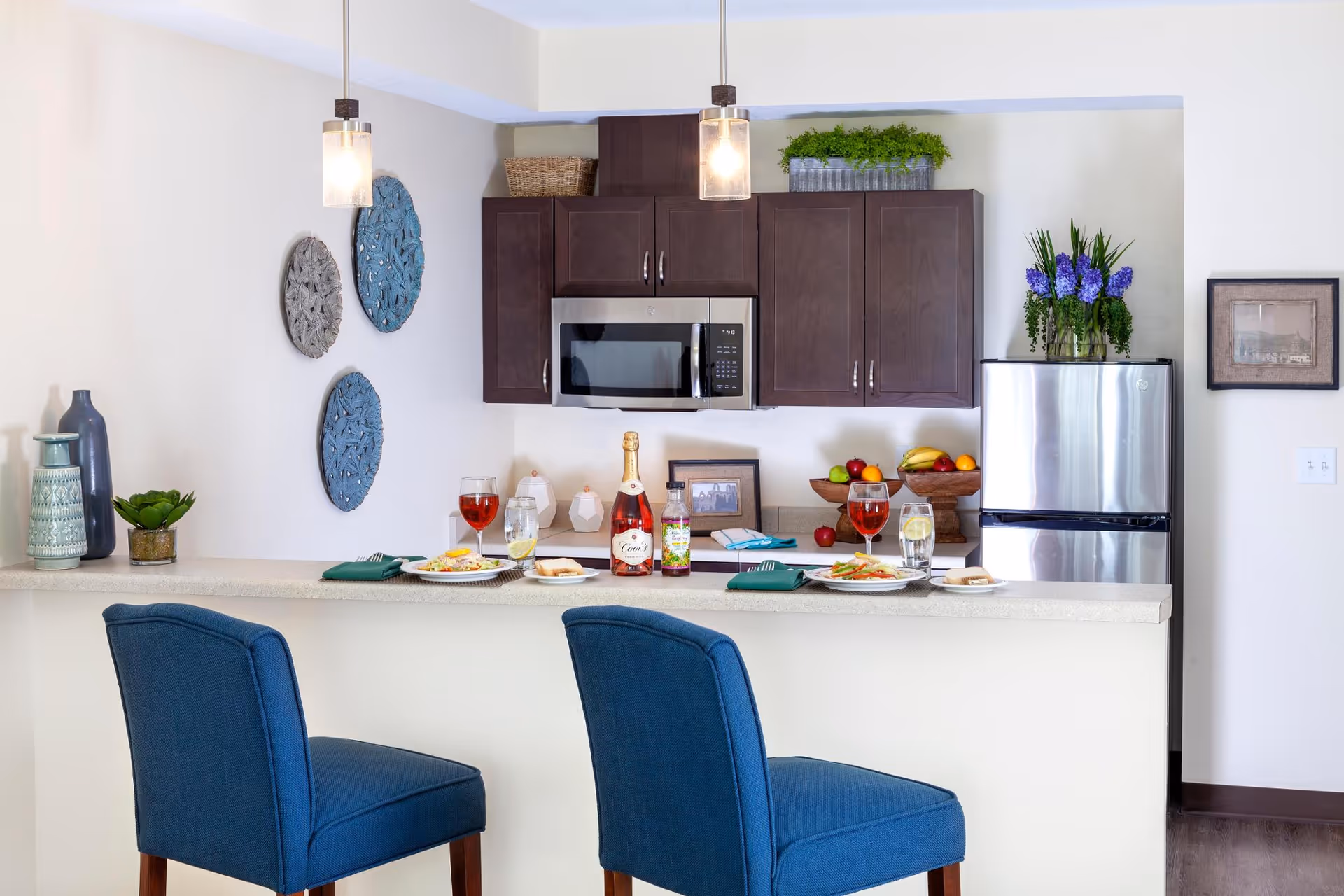 Open kitchen with a breakfast bar, two blue bar stools, dark cabinets, stainless microwave and refrigerator, and place settings on the counter.