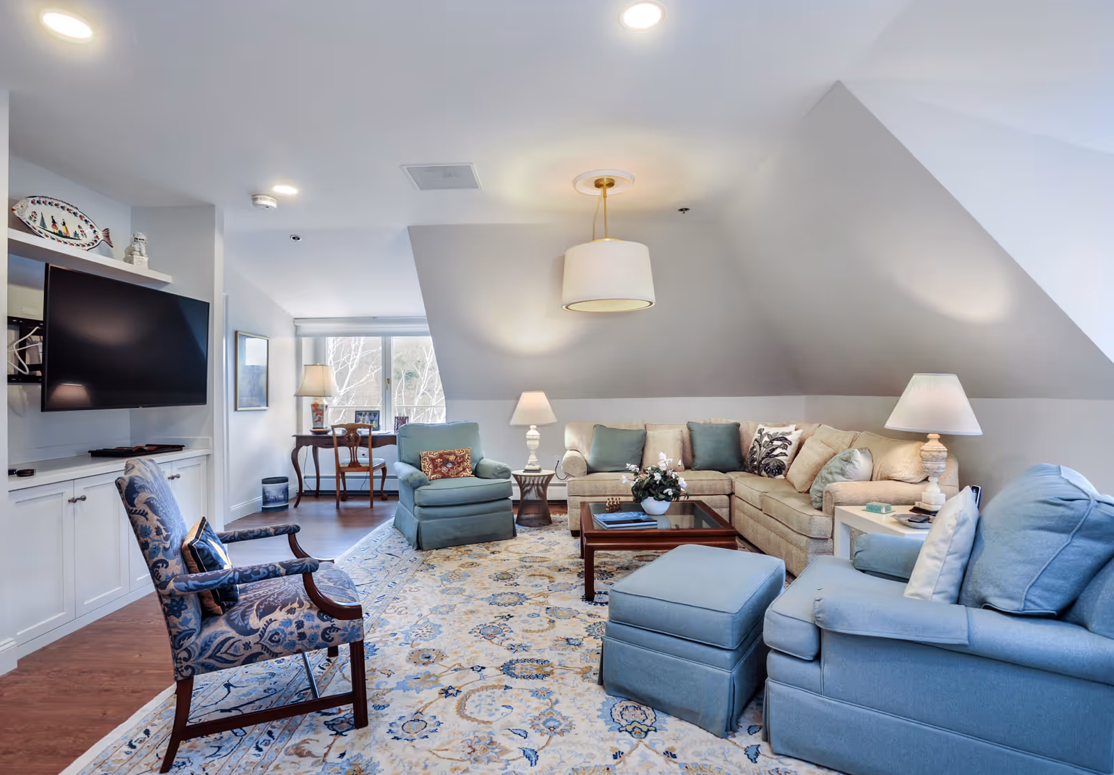 Spacious, well-lit living room with a beige sectional, blue armchairs and ottoman, patterned rug, coffee table and wall-mounted TV.