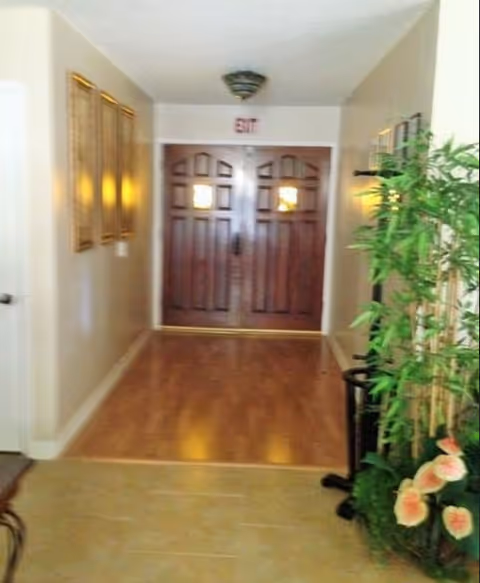 Interior hallway with wood floors leading to double wooden exit doors, framed pictures on the left and plants on the right.