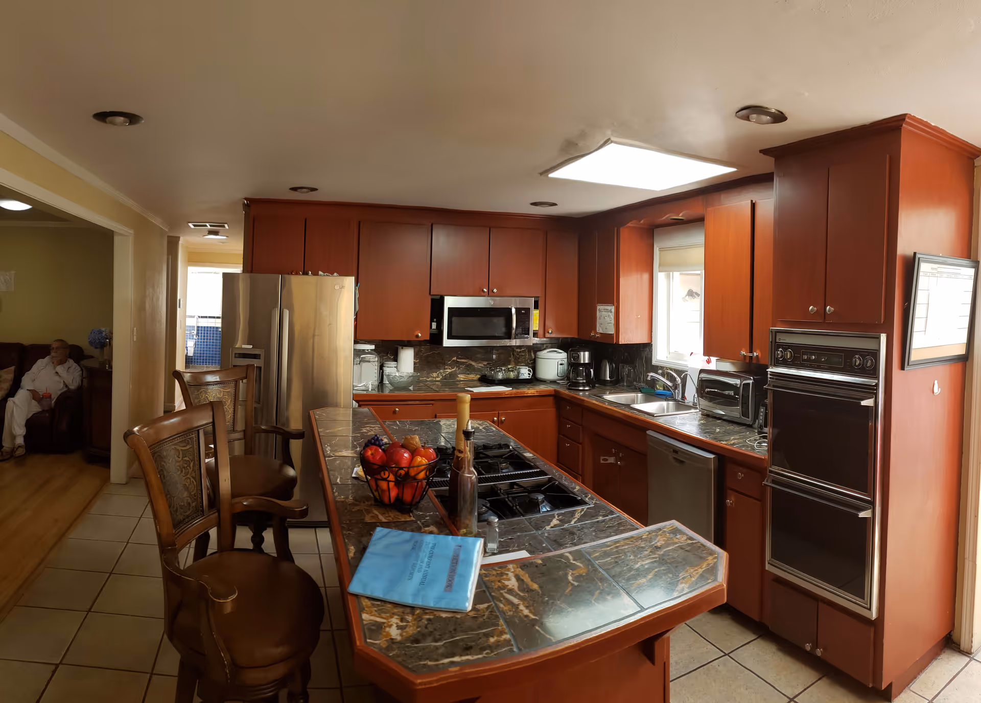 A kitchen with dark marble countertops and wooden cabinets. The kitchen features a stainless steel refrigerator, built-in microwave, double oven, dishwasher, and various small appliances on the counters. There are two wooden chairs at the kitchen island, which has a fruit basket and a blue booklet on it. In the background, a person is sitting in a living room area visible through a doorway.