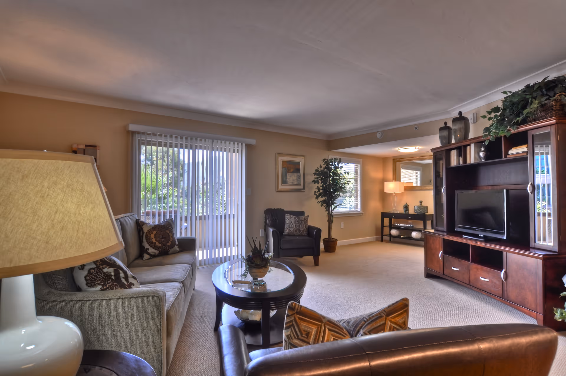A well-lit living room with a beige sofa, a dark leather armchair, a round glass coffee table, and a large wooden entertainment center with a TV. There is a lamp on a side table, a potted plant near the window, and vertical blinds covering a sliding glass door leading outside.