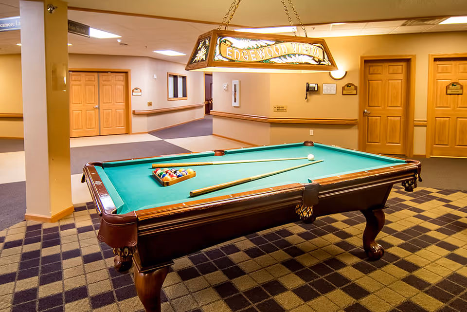 Interior view of a senior living facility game room featuring a pool table with billiard balls and cues set up. The room has beige walls, wooden doors, and a patterned carpet. A hanging light fixture above the pool table displays the text 'EDGEWOOD VISTA'.