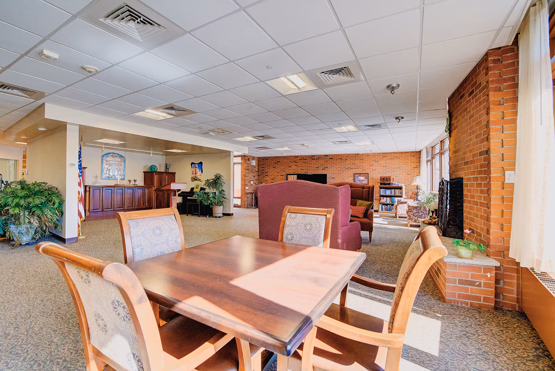 A bright and spacious common area in a senior living facility with a wooden table and four upholstered chairs in the foreground. In the background, there are comfortable armchairs, a bookshelf, a piano, and decorative plants. The room features large windows with curtains, brick walls, and a carpeted floor.
