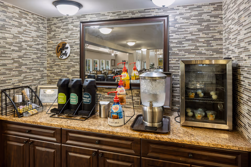 A beverage station with three coffee dispensers labeled Holiday Hot Water, Holiday Decaf Coffee, and Holiday Regular Coffee, a water dispenser filled with ice water, and a small refrigerated display case containing cups of fruit. The station is set against a wall with mosaic tile backsplash and a large mirror reflecting a dining area with tables and chairs.